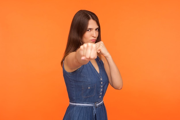 Chica morena decidida con espíritu de lucha en vestido casual golpeando con puños boxeando mirando con confianza a la cámara mujer fuerte defendiéndose tiro de estudio aislado sobre fondo naranja