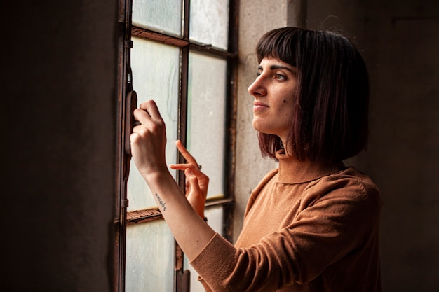 Chica morena delante de una ventana mirando por la ventana, imagen con poca luz