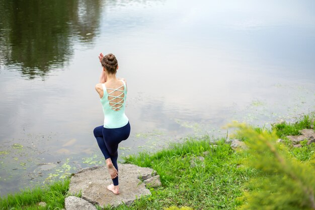 Chica morena delgada haciendo yoga en el verano en un césped verde junto al lago