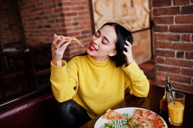 Chica morena divertida en suéter amarillo comiendo pizza en el restaurante