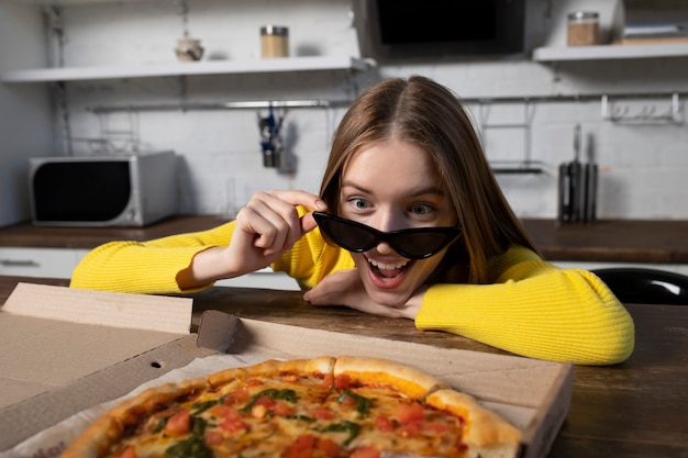 Chica morena divertida en suéter amarillo y gafas negras comiendo pizza en la cocina