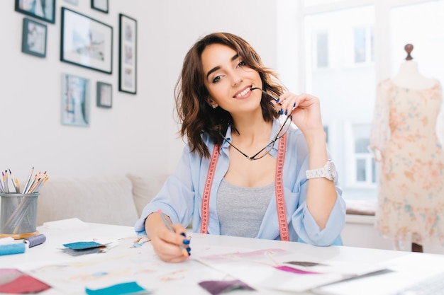 Una chica morena está sentada a la mesa en el taller. Ella tiene una camisa azul y un desorden creativo en la mesa. Tiene gafas y manos de lápiz y sonríe a la cámara.