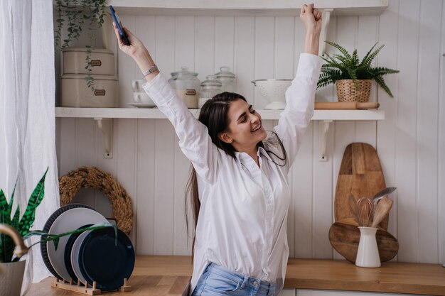 Chica morena feliz en la cocina vestida con camisa blanca y jeans azules levantó las manos sonriendo ampliamente