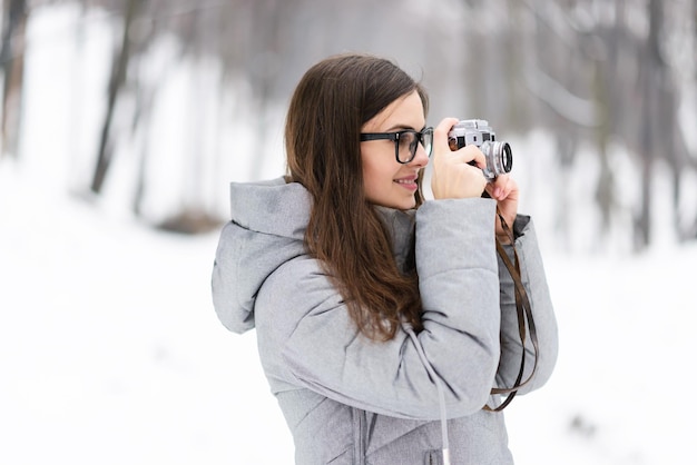 Chica morena con gafas y chaqueta gris fotografiando paisajes invernales mientras camina por el bosque