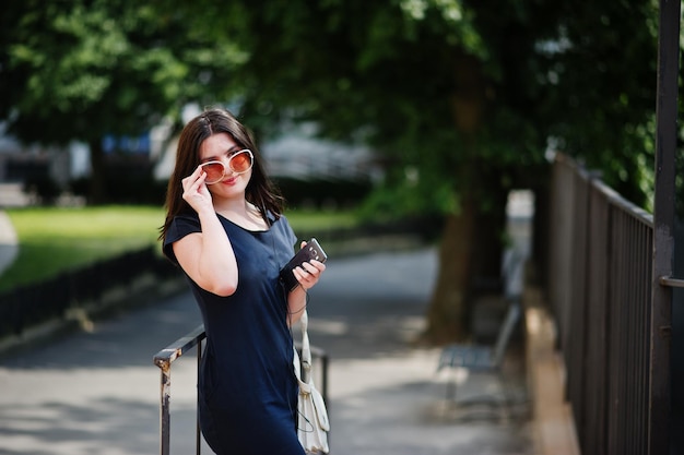 Chica morena con gafas de sol de vestido negro escuchando música desde el teléfono de los auriculares y posando en la calle de la ciudad