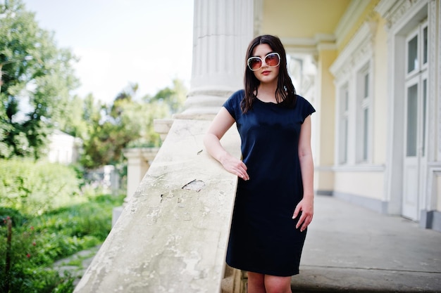 Chica morena con gafas de sol de vestido negro posadas contra la antigua casa vintage en la calle de la ciudad