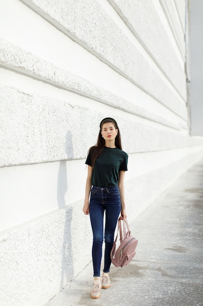 Chica morena en jeans, una camiseta verde, una banda para el cabello y una mochila rosa se alza contra una pared blanca