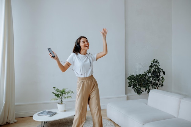 Chica morena joven feliz en una camiseta blanca y jeans beige en casa escuchando música en los auriculares