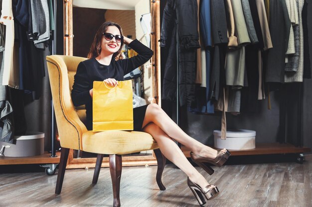 Chica morena joven de moda con gafas de sol sentada en el sillón amarillo sosteniendo una bolsa de compras amarilla en el concepto de viernes negro del centro comercial