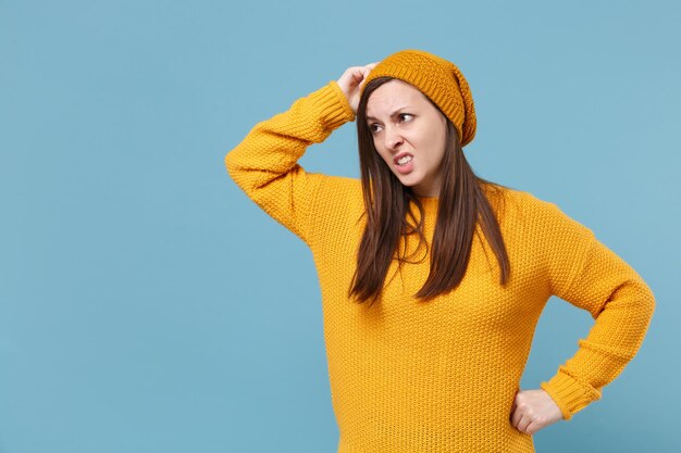 Chica morena joven preocupada con sombrero de suéter amarillo posando aislada en un retrato de estudio de fondo azul. Gente emociones sinceras concepto de estilo de vida. Simulacros de espacio de copia. Ponerse la mano en la cabeza.