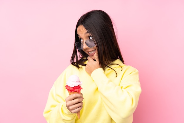 Chica morena joven sosteniendo un helado de cucurucho sobre fondo rosa aislado mirando hacia el lado