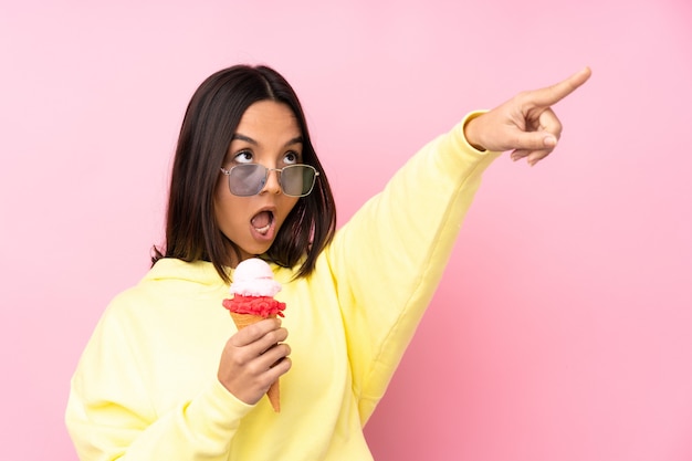 Chica morena joven sosteniendo un helado de cucurucho sobre pared rosa aislada apuntando hacia afuera