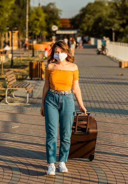 Chica morena con una mascarilla quirúrgica camina en el parque con su maleta.