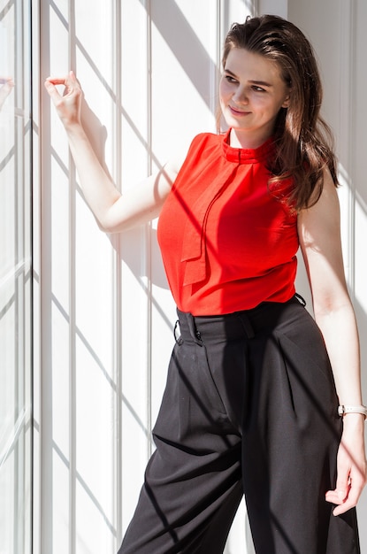 Chica morena de moda en una blusa roja posa cerca de la ventana y disfruta de la luz del sol y el calor.