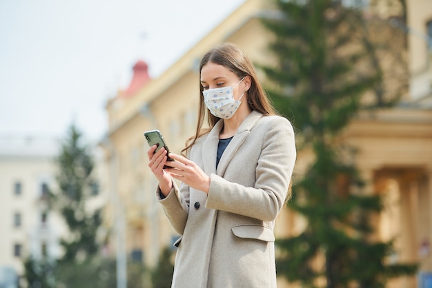 Una chica morena con el pelo largo y una máscara médica para evitar la propagación del coronavirus lee las noticias en un teléfono inteligente en la calle vieja. Una mujer con una máscara facial contra COVID-19 mantiene una distancia social.