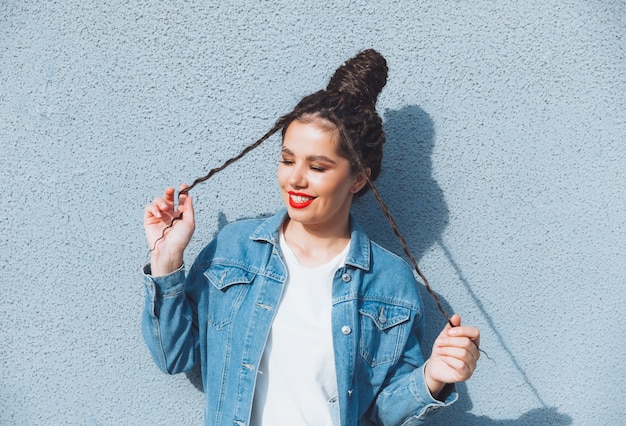 Una chica morena con rastas y lápiz labial rojo en un traje de mezclilla está parada cerca de una pared azul generación z