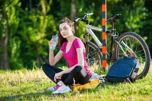 Chica morena en ropa deportiva con una bicicleta Descansando y bebiendo agua