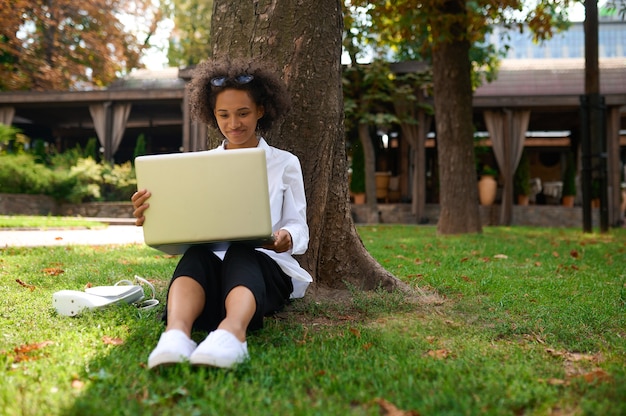 Chica morena sentada bajo el árbol con una computadora portátil