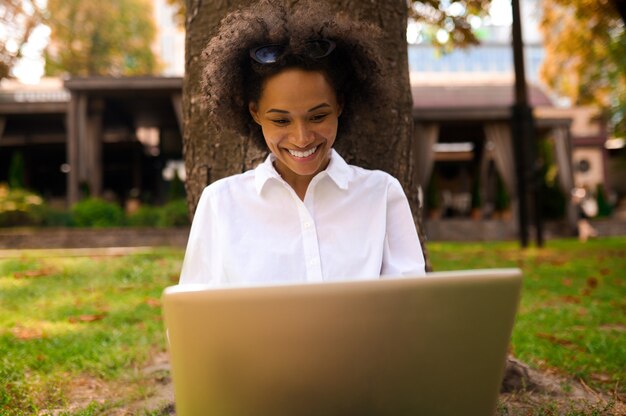 Chica morena sentada bajo el árbol con una computadora portátil