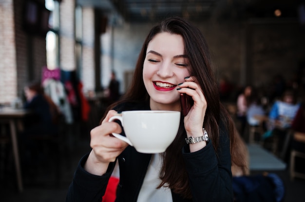 Chica morena sentada en la cafetería con una taza de capuchino y hablando de teléfono móvil.