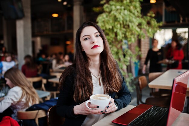 Chica morena sentada en la cafetería con una taza de capuchino, trabajando con ordenador portátil rojo.