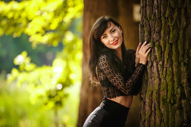 Chica morena sexy en una blusa de encaje y falda de cuero con tacones altos posando cerca de un árbol en el bosque