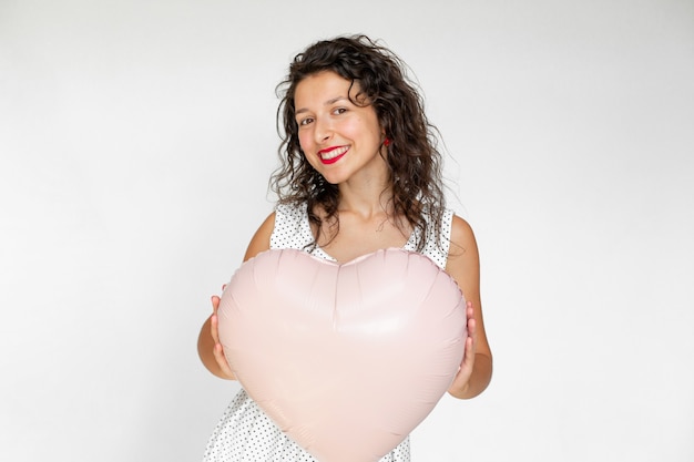 Chica morena sexy posando con globos en forma de corazón sobre un fondo blanco.