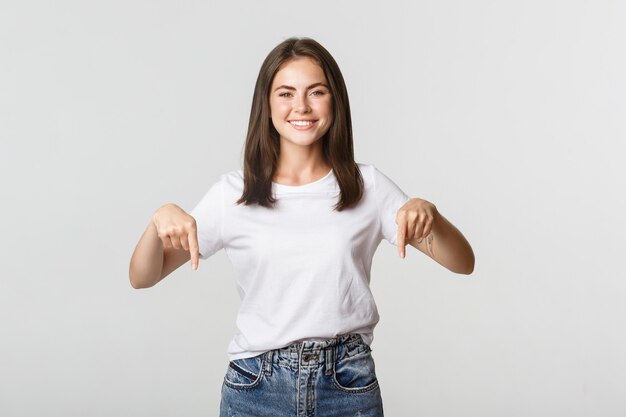 Chica morena sonriente satisfecha apuntando con el dedo hacia abajo, recomendar producto.