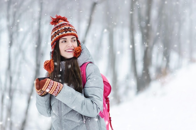 Chica morena turista con chaqueta gris, sombrero y mitones de pie con taza termo y considerando los paisajes invernales en el bosque frío de invierno