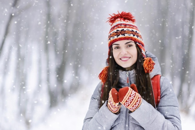 Chica morena turista con chaqueta gris, sombrero y mitones sonriendo y sosteniendo una taza termo en el bosque frío de invierno