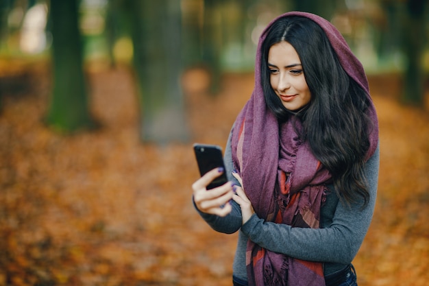 chica morena usando su teléfono mientras camina por el parque durante el otoño