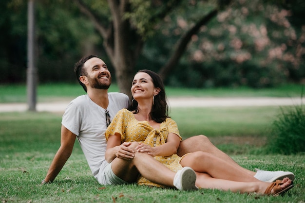 Una chica morena con un vestido amarillo está sentada entre las piernas de su novio en el césped del casco antiguo de España. Un hombre está abrazando a su novia en una cita en el parque valenciano.