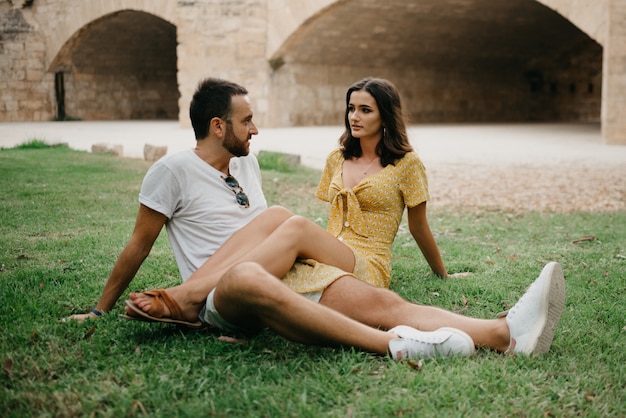 Una chica morena con un vestido amarillo está sentada entre las piernas de su novio en el césped en el casco antiguo de España. Una pareja de turistas en una cita en el parque valenciano.