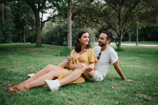 Una chica morena con un vestido amarillo está sentada entre las piernas de su novio en el césped en el casco antiguo de España. Una pareja de turistas riendo en una cita en el parque valenciano.