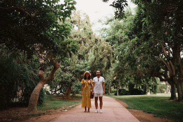 Una chica morena con un vestido amarillo y su novio se alojan en un camino de arena entre árboles en el parque valenciano. Un par de turistas en una cita en verano por la noche.