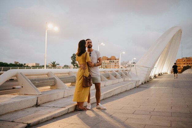 Una chica morena con un vestido amarillo y su novio sonriente se abrazan en un puente blanco en Valencia. Un par de turistas de cuerpo entero en una cita en la cálida noche.