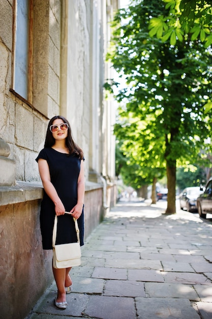 Chica morena en vestido negro con gafas de sol con bolso a mano posando en la calle de la ciudad