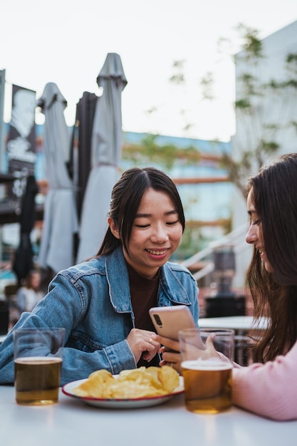 Una chica mostrando algo a su amiga en un teléfono móvil en la terraza de un bar Están tomando una cerveza