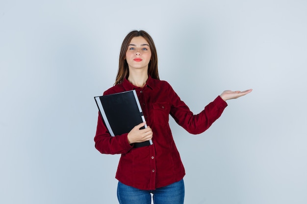 chica mostrando gesto de bienvenida, sosteniendo una carpeta en camisa burdeos y luciendo confiada.