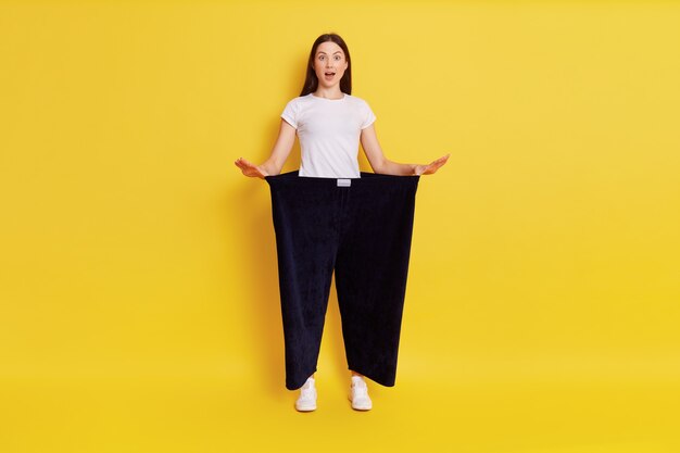 Chica mostrando pérdida de peso, chica asombrada con la boca abierta, dama de camiseta blanca con pantalones negros viejos de gran tamaño, de pie aislado sobre una pared amarilla.