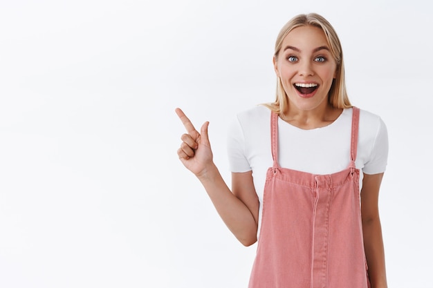 Chica mostrando promo con emoción y alegría. Atractiva mujer rubia con un mono rosa, camiseta apuntando hacia la esquina superior izquierda, con la boca abierta fascinada, sonriendo impresionado y mirando a la cámara, fondo blanco