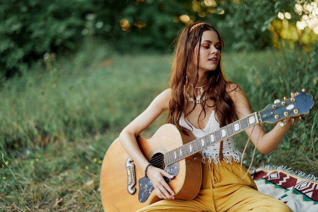 Chica mujer hippie tocando la guitarra con ropa ecológica sentada en el suelo afuera en la naturaleza en otoño viendo la puesta de sol