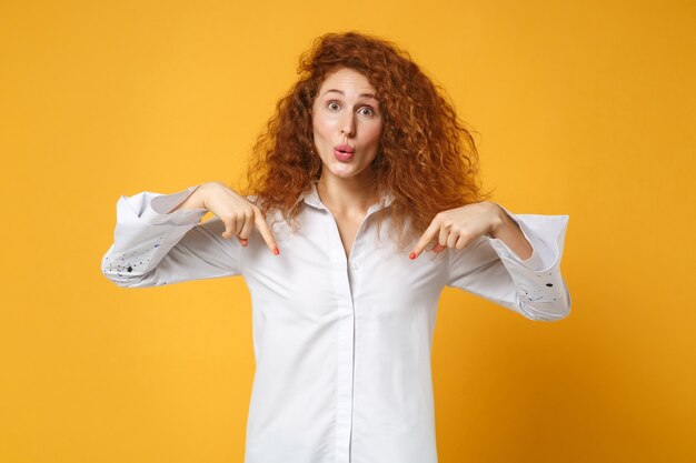 Chica de mujer pelirroja joven sorprendida en camisa blanca posando aislada en la pared naranja amarilla