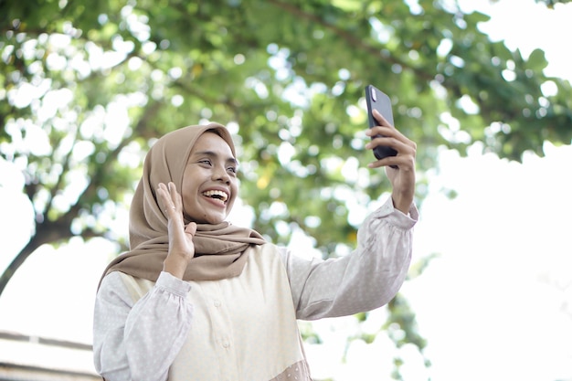 Chica musulmana asiática con ropa informal está usando un teléfono inteligente y sonriendo mientras está de pie en el área al aire libre