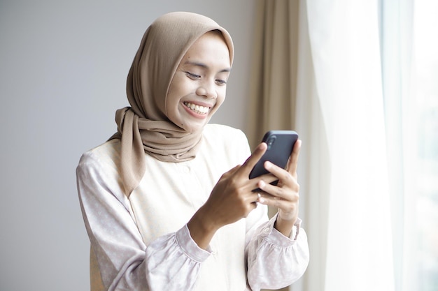 Chica musulmana asiática con ropa informal está usando un teléfono inteligente y sonriendo mientras está de pie cerca de la ventana