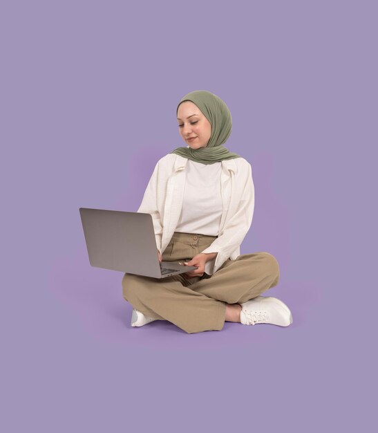 Chica musulmana estudiante universitaria caucásica en hiyab usando laptop Vista de cuerpo completo sentarse en el piso