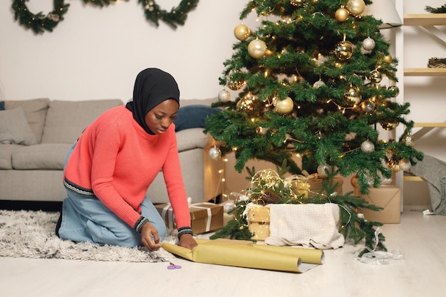 Chica musulmana en hiyab negro de pie cerca de la caja de regalo de envoltura de árbol de Navidad