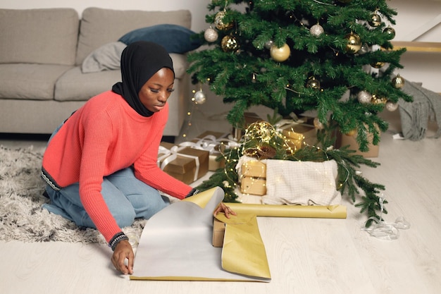 Chica musulmana en hiyab negro de pie cerca de la caja de regalo de envoltura de árbol de Navidad