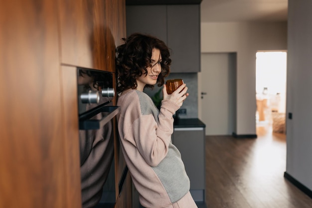 Chica muy encantadora con rizos en traje de casa bebiendo té en la cocina y descansando