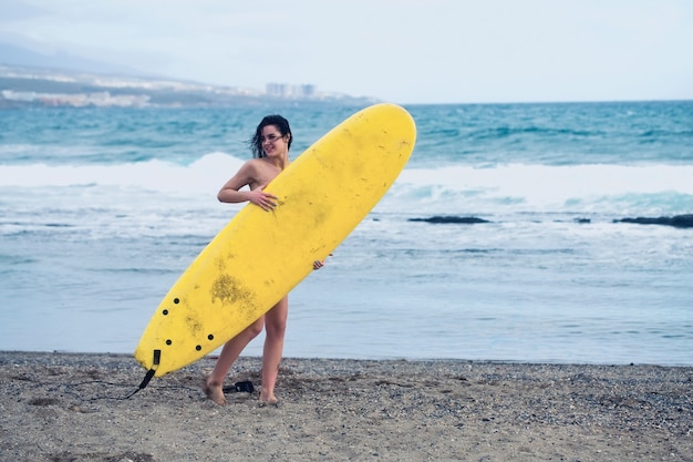 Chica muy feliz, surfista o mujer hermosa, morena delgada sexy, en traje de baño sexi con tabla de surf amarilla en la playa de arena al aire libre en un día soleado de verano en el fondo del mar azul